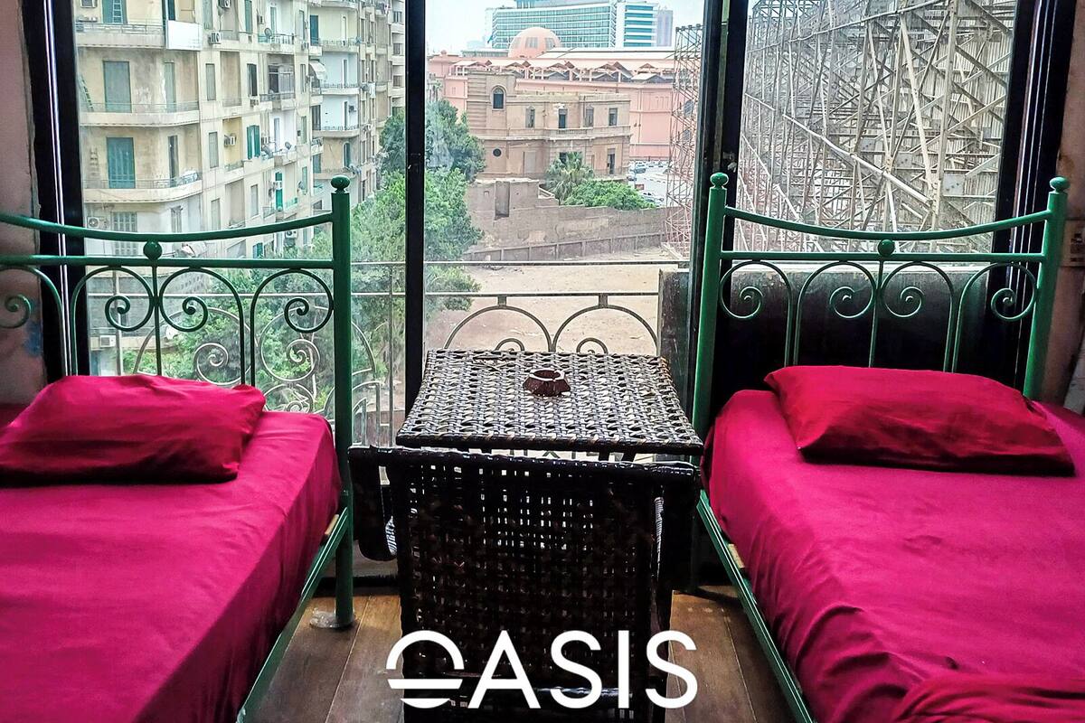 Habitación con dos camas, una mesita y vistas a edificios y una noria a través de amplios ventanales.