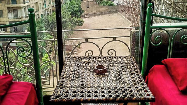 A balcony with a woven table and red cushions, overlooking a cityscape with buildings and a construction site. - OASIS Hotel Downtown (Cairo)