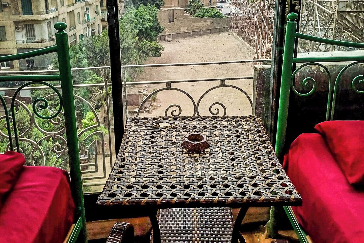 A balcony with a woven table and red cushions, overlooking a cityscape with buildings and a construction site.