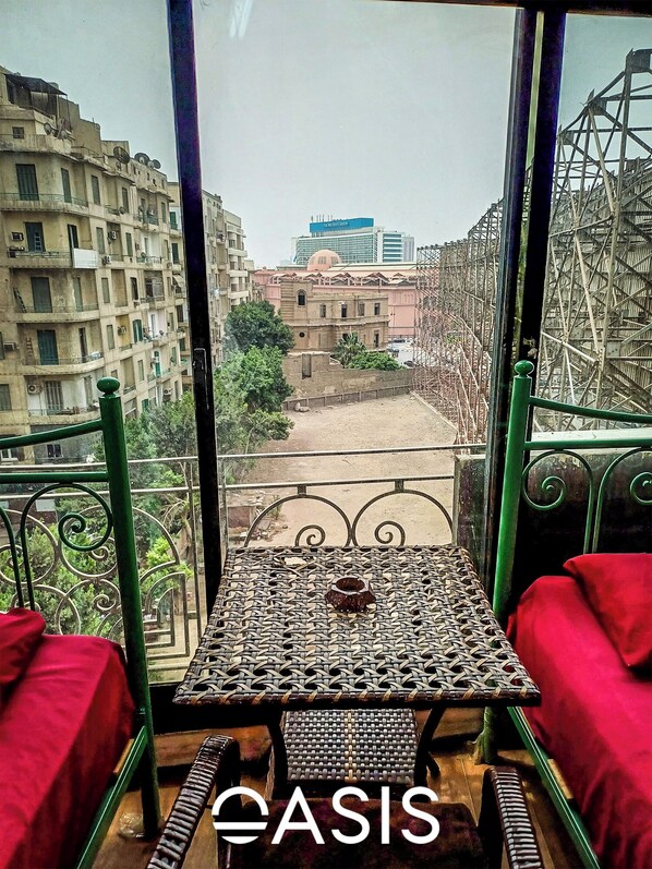 A balcony with a woven table and red cushions, overlooking a cityscape with buildings and a construction site. - OASIS Hotel Downtown (Cairo)