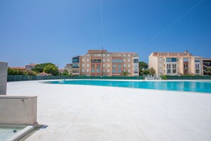 Une piscine avec une terrasse carrelée, un bâtiment doté de nombreuses fenêtres et de balcons, et un ciel bleu dégagé.