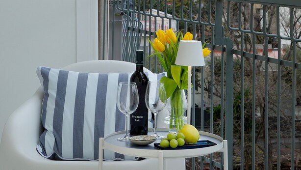 A balcony with a white chair, a striped pillow, a small table with a bottle, a glass, a vase with yellow flowers, and a view of a tall building with a cross.