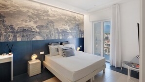 A modern bedroom with a large bed, a nightstand, a wall-mounted lamp, and a view of the city through a balcony.