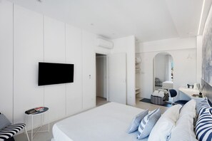 Una habitación de hotel moderna con una cama grande, un televisor de pantalla plana montado en la pared, una mesita auxiliar y una zona de estar con un banco y cojines.