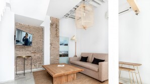 A modern living room with a wooden coffee table, a sofa, a TV mounted on a brick wall, and a hanging light fixture. - Superior Suite - Open Space (Sevilla)