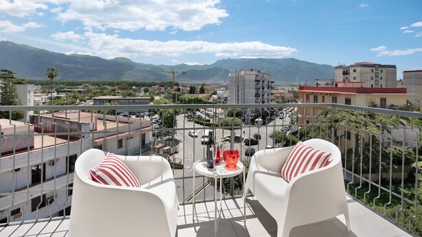 Ein Balkon mit weißen Stühlen und einem kleinen Tisch, mit Blick auf eine Stadtlandschaft und dahinter Berge.