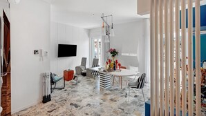Ein modernes Wohnzimmer mit Steinboden, einem Flachbildfernseher, einer Essecke mit gestreiften Stühlen und einem weißen Vorhang.