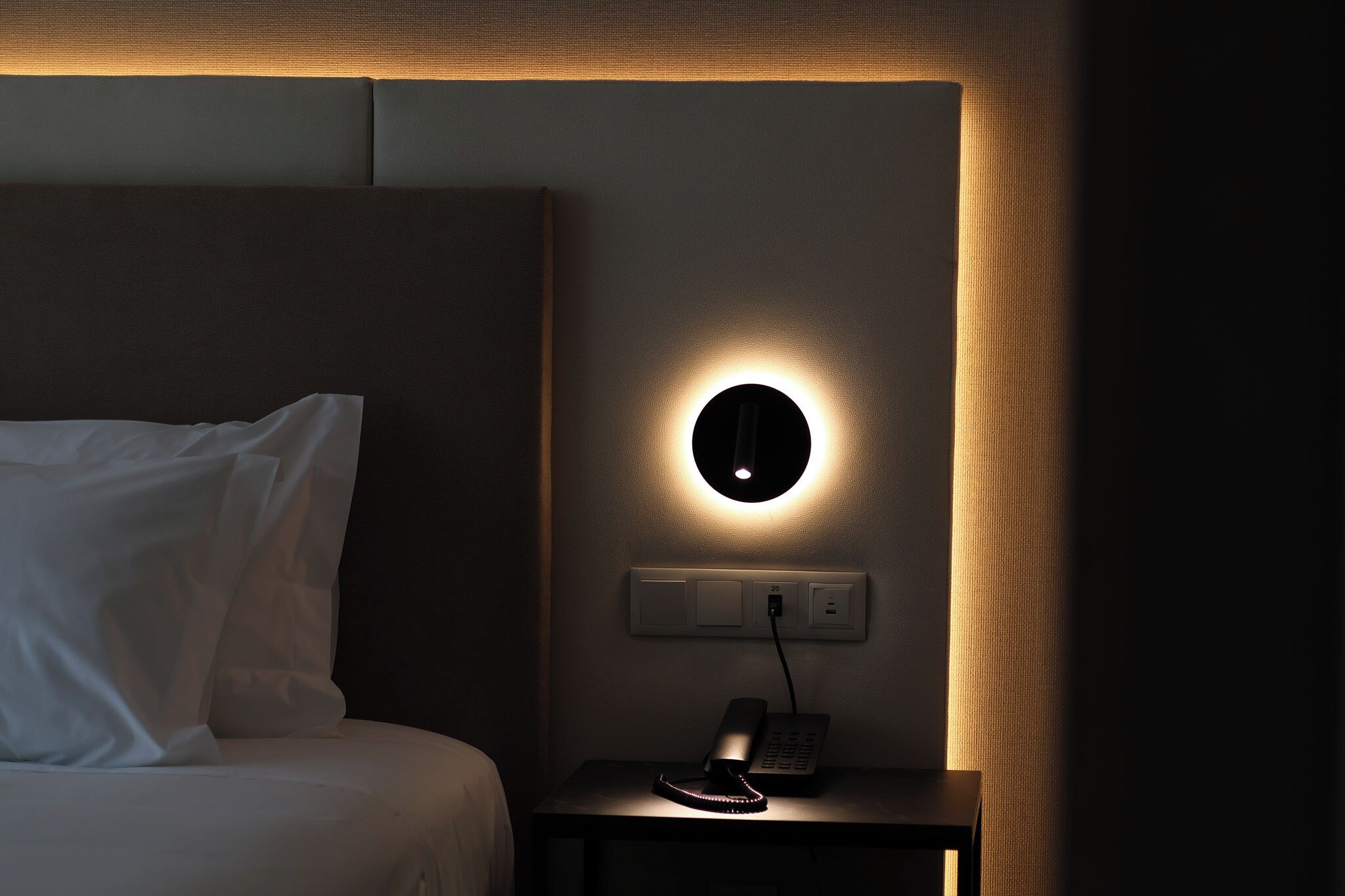 A hotel room with a bed, a bedside table with a phone, and a wall-mounted light fixture.