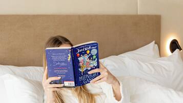 Una persona recostada en una cama con ropa de cama blanca, sosteniendo un libro titulado "Midnight Games".