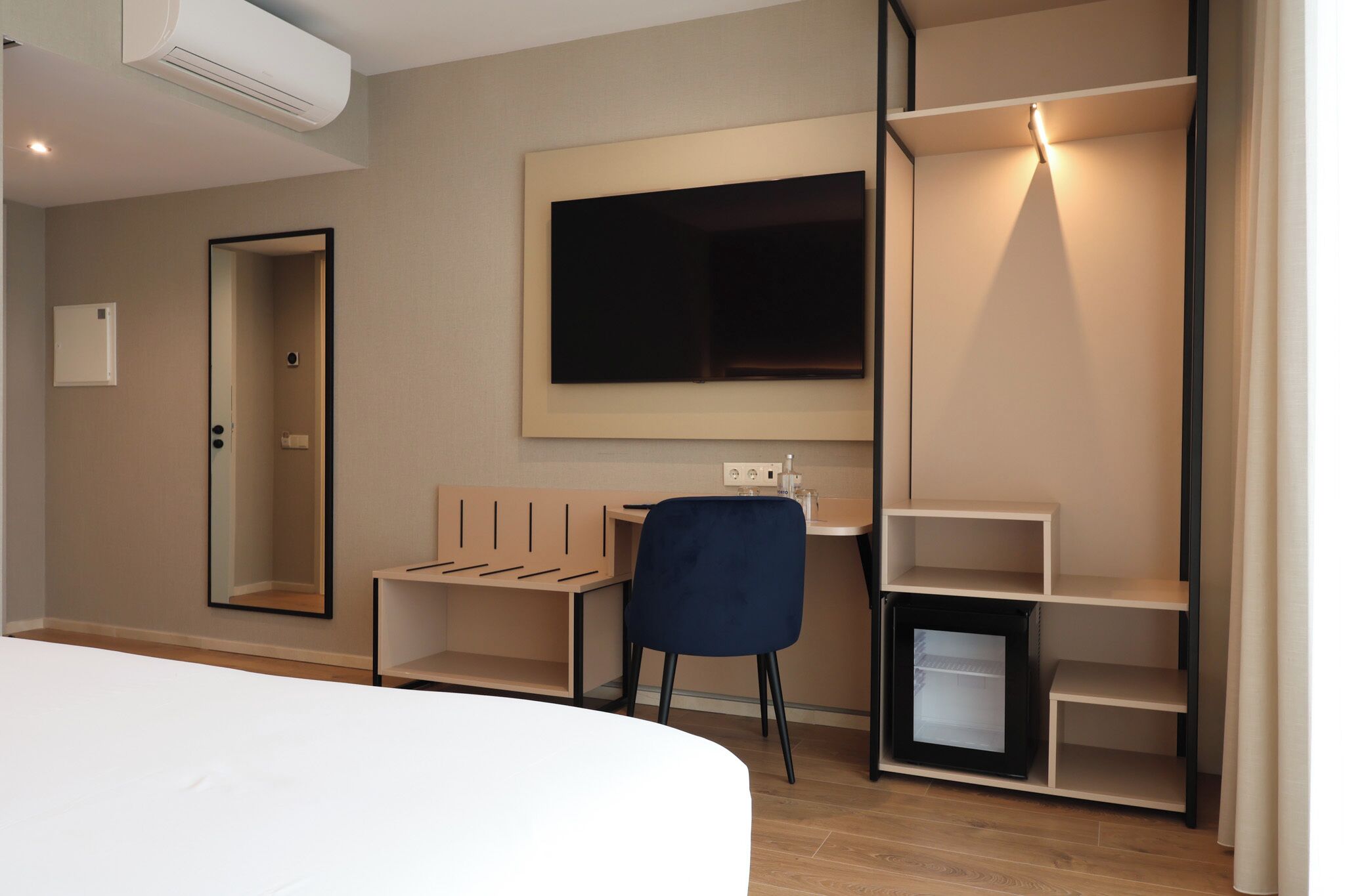 A modern hotel room with a bed, a desk, a chair, a TV, and built-in shelving.