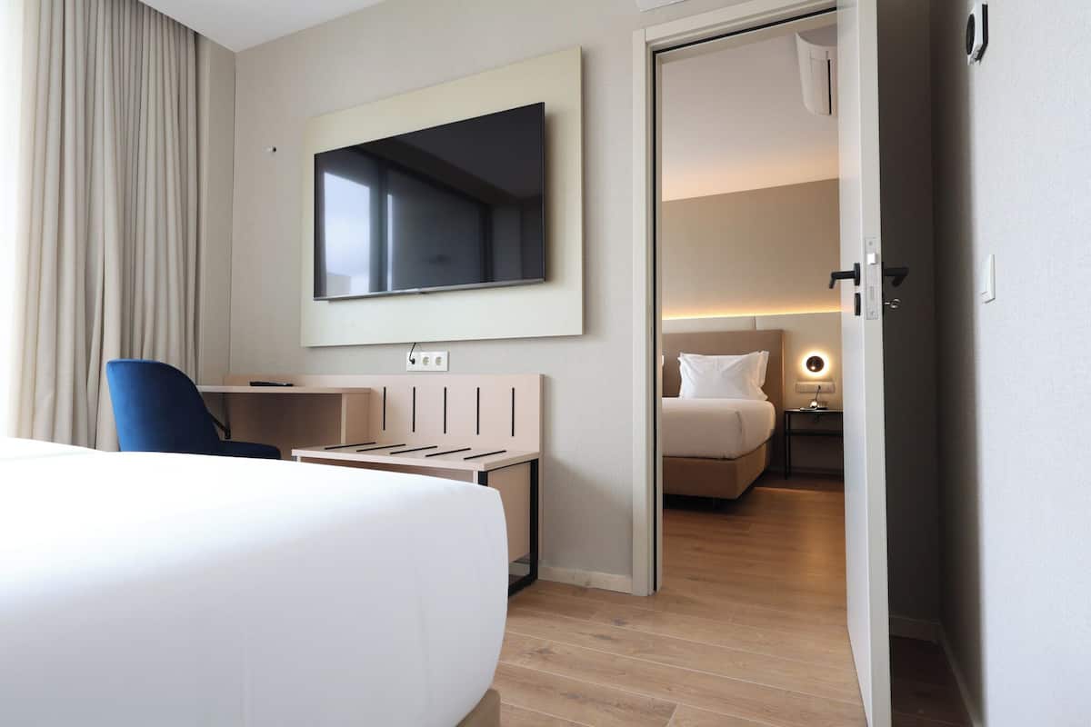 A hotel room with a bed, a desk, a chair, a TV mounted on the wall, and a separate bedroom with a bed and nightstand.