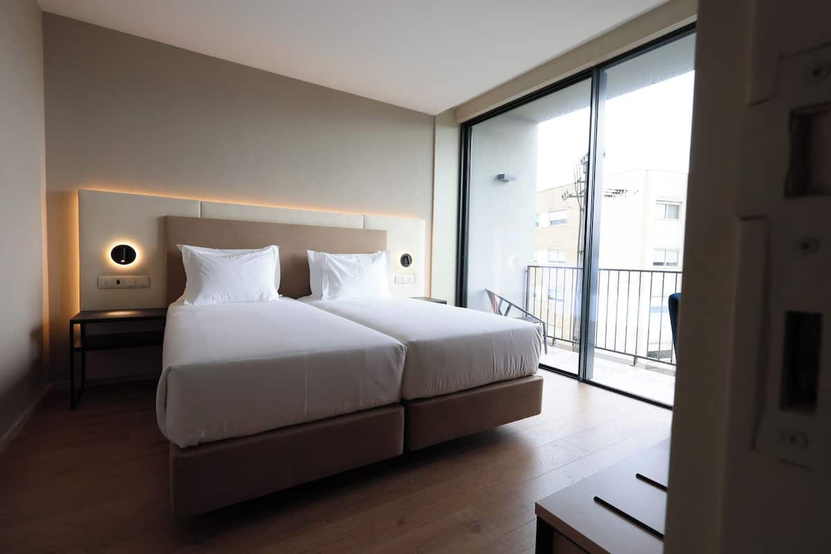 A modern hotel room with a large bed, a bedside table with a lamp, and a view of the outside balcony.