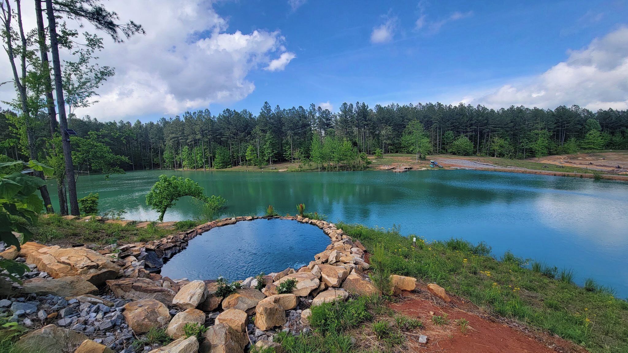 Paisagem natural com lago, cercado por árvores e uma área rochosa com uma pequena piscina.