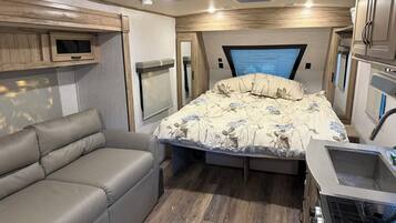 Interior compacto de una autocaravana con cama, sofá, kitchenette y televisor.