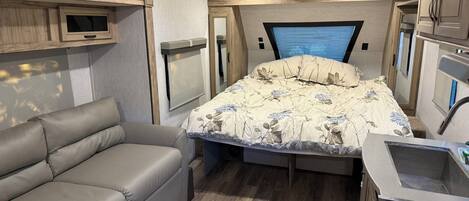 Interior compacto de una autocaravana con cama, sofá, kitchenette y televisor.