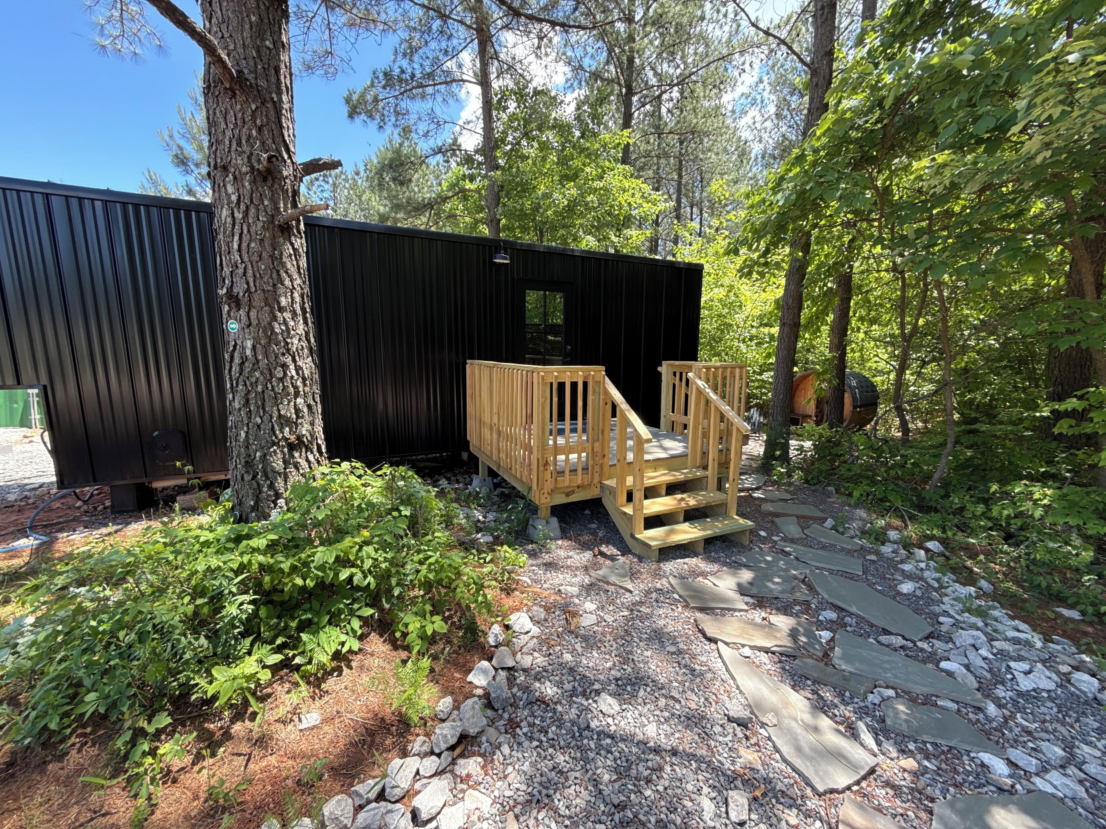 A wooden deck with a staircase leading to a black metal structure, surrounded by trees and gravel.