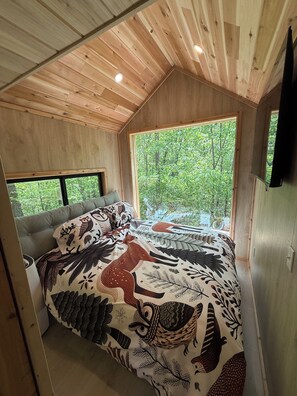 A cozy bedroom with a large bed, a window with a view of greenery, and wooden ceiling beams. - Sasquatch Farm (South Pittsburg)