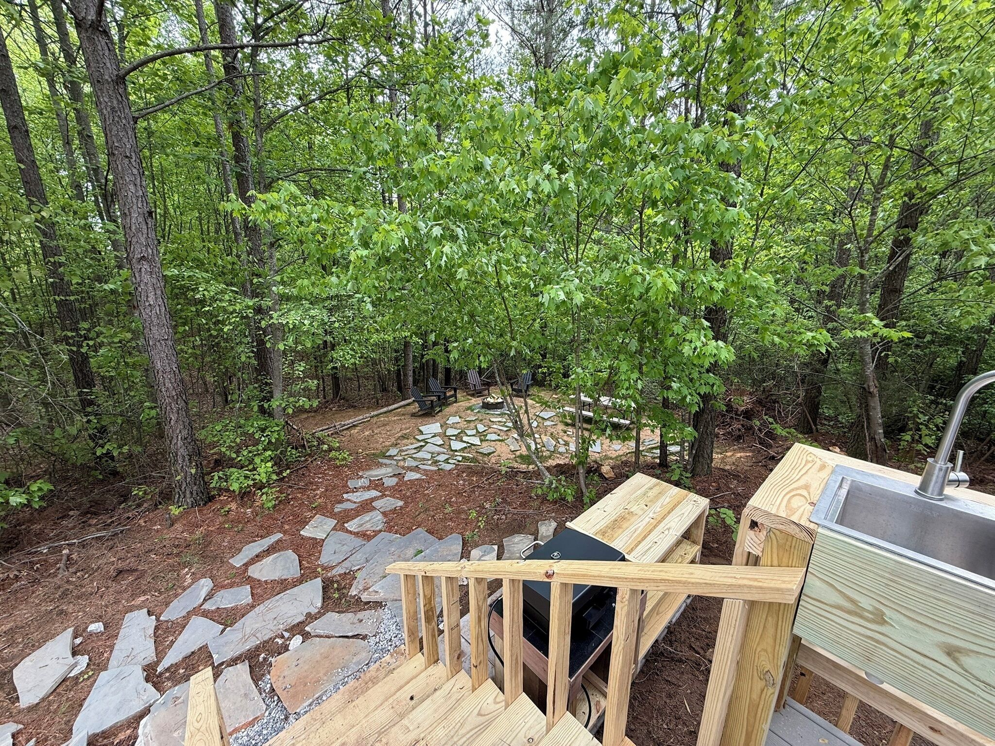 A wooden deck with a stone pathway leading to a seating area surrounded by trees.