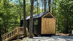 A cabin with a wooden exterior, a staircase leading to a deck, and a gravel driveway surrounded by trees.