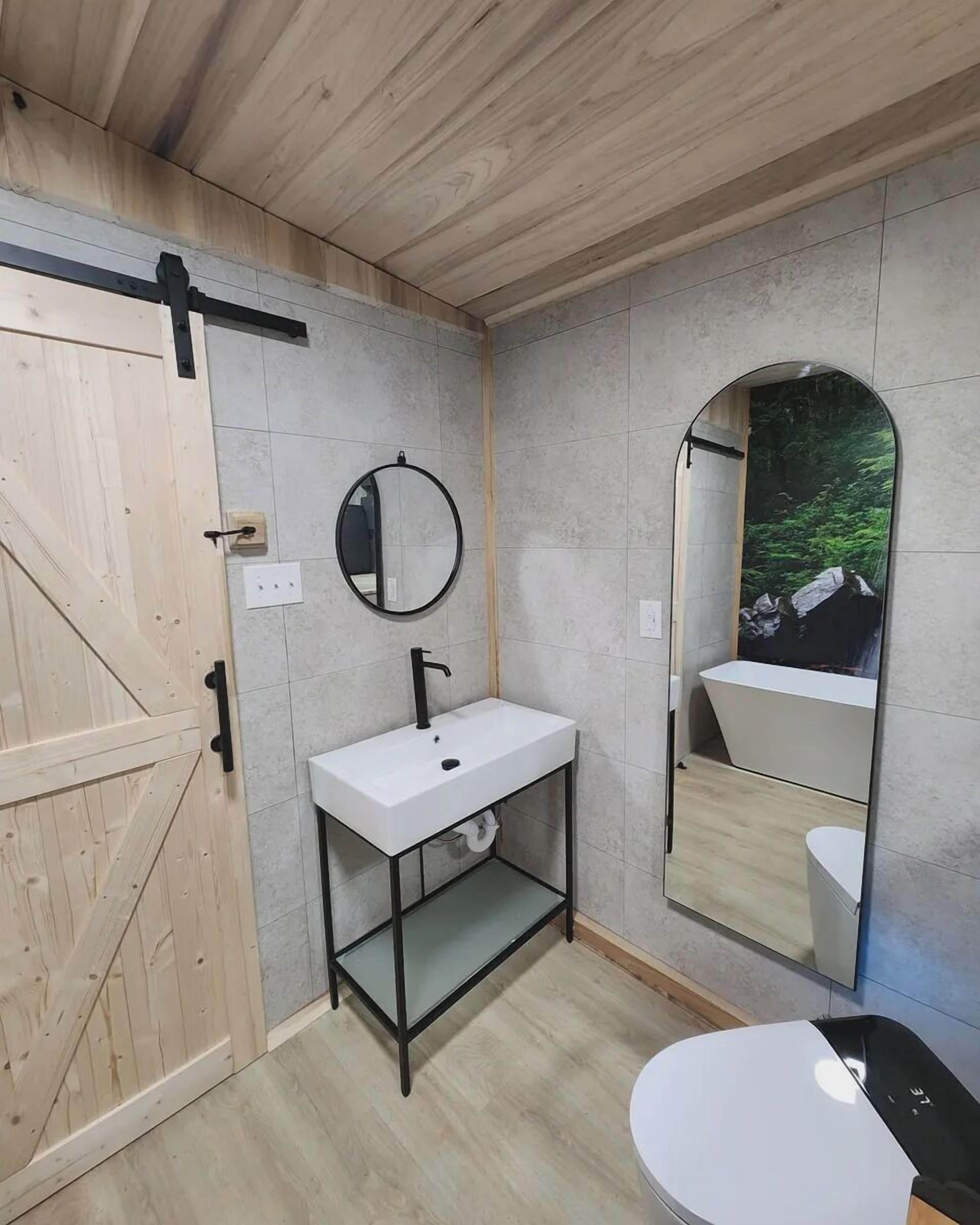 A modern bathroom with a wooden door, a white sink with a black faucet, a round mirror, and a large mirror reflecting a forest scene.