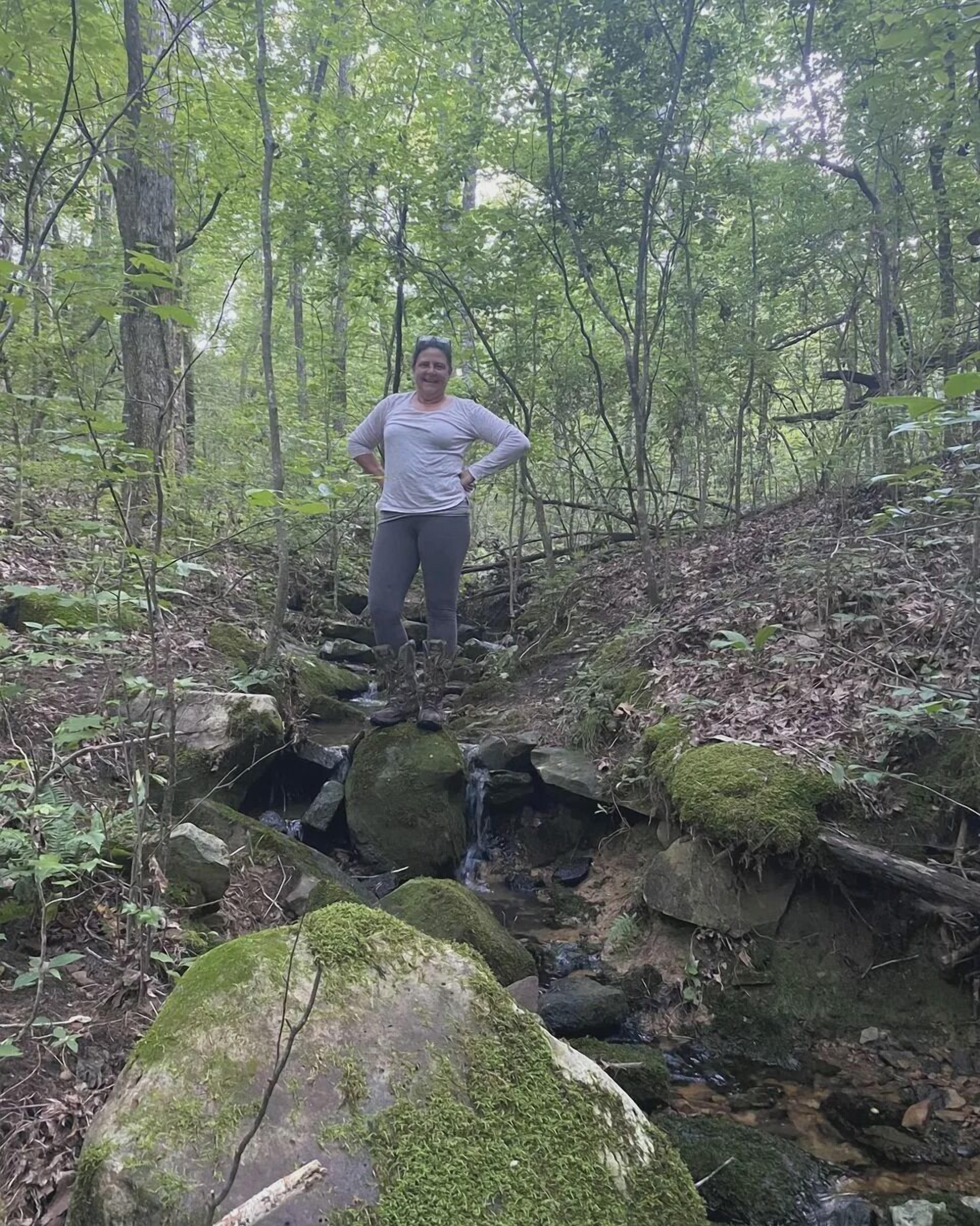 A person standing on a moss-covered rock bridge in a forested area with a stream below.