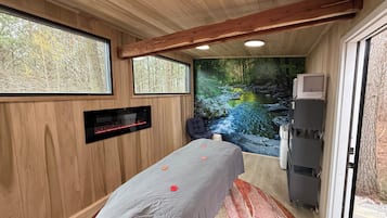 Una habitación de madera con una cama, una camilla de masaje, un televisor y un mural en la pared que representa un arroyo.