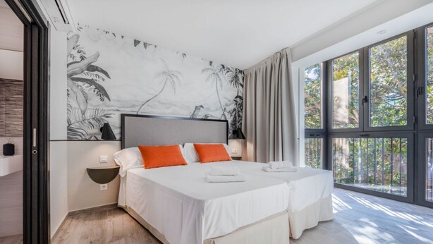 A modern hotel room with a large bed, two pillows, a nightstand, a wall-mounted lamp, and a large window with a view of trees.