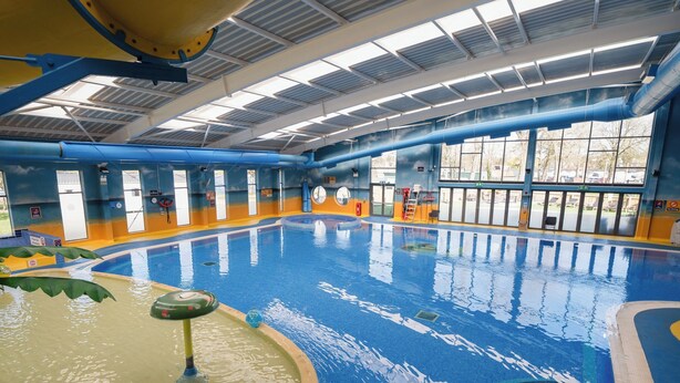 Ein Hallenschwimmbad mit glatter Wasseroberfläche, Sprungbrett und Rettungsschwimmerstuhl.
