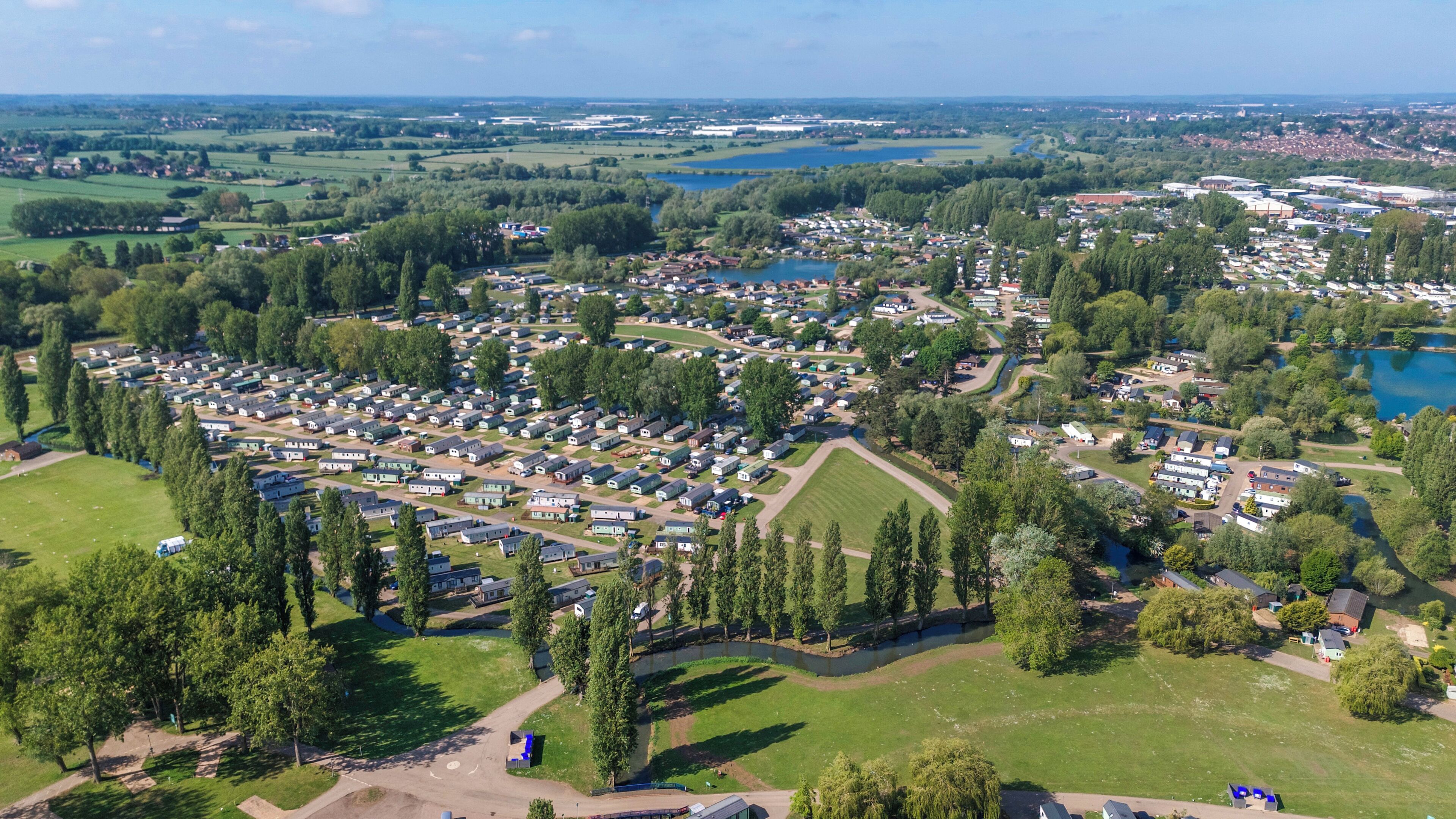 Eine Vogelperspektive zeigt einen Campingplatz mit zahlreichen Wohnmobilen und Wohnwagen, umgeben von grünen Feldern und einem See.