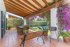 An outdoor patio with a wooden bench, a table with chairs, a grill, and potted plants.