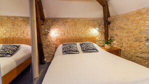 A bedroom with a stone wall, wooden beams, a bed with patterned pillows, and a bedside table with a plant.