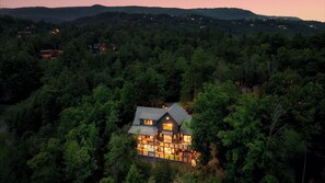 A house surrounded by dense forest with multiple windows illuminated, indicating interior lighting.