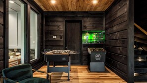 A room with a foosball table, a television displaying a video game, and a cozy seating area with a chair. - Escape in Luxury •  Pool, HotTub, Sauna & GameRoom (Sevierville)