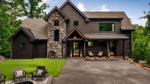A two-story house with a stone tower, a covered porch with seating, and a well-maintained lawn. - Escape in Luxury •  Pool, HotTub, Sauna & GameRoom (Sevierville)