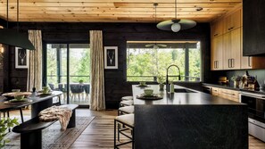 A modern kitchen with a central island, wooden cabinetry, and large windows offering a view of greenery.