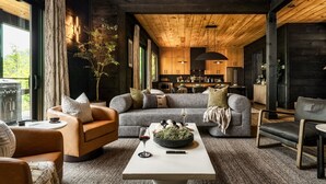Un salon moderne avec un canapé gris, une table basse sur laquelle se trouvent un saladier et des verres à vin, et une vue sur la cuisine et l’espace repas grâce à de grandes fenêtres.