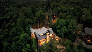 A large, dark-colored house surrounded by dense forest, with multiple illuminated windows and a visible outdoor area.