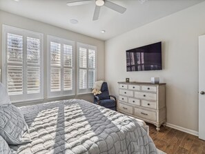Ein Schlafzimmer mit Bett, Kommode, Stuhl, Deckenventilator und einem Fenster mit Jalousien.
