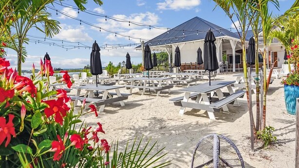 A beachside patio with white tables and black umbrellas, a covered seating area, and a building with a dark roof.