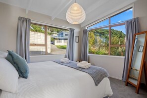 A bedroom with a large bed, a standing lamp, and a view of greenery outside. - Tideview Cottage - Tairua (Tairua)