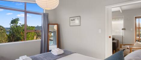 Un dormitorio con un ventanal grande, una cama, una mesita de noche, una lámpara colgante y una puerta que da a otra habitación.