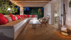 An outdoor patio with a long white sofa, red cushions, wicker chairs, a table, and string lights.