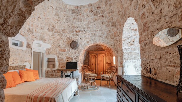 A rustic stone-walled bedroom with a bed, two wicker chairs, a wooden cabinet, and a television.