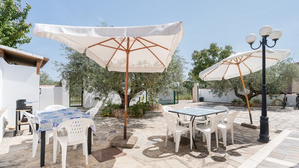 An outdoor patio with stone flooring, white plastic chairs, a table with a blue patterned tablecloth, and two large white umbrellas. - Apartment 'Sa Curcurica' (Cala Liberotto)
