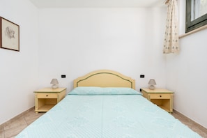 Ein Schlafzimmer mit einem Bett, zwei Nachttischen mit Lampen, einem gerahmten Bild an der Wand und einem Fenster mit Vorhängen.