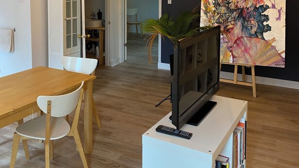 A modern living room with a wooden dining table, a TV on a white stand, a bookshelf, and a large abstract painting on the wall. - Apartment for rent in Bromont - 1 bedroom 1 bathroom (Bromont)