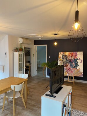 Una sala de estar moderna con una mesa de comedor de madera, un televisor sobre un mueble blanco, una estantería y un cuadro abstracto de gran tamaño en la pared.