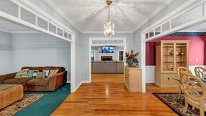 A living room with a wooden floor, a brown sofa, a coffee table, a chandelier, a TV, a fireplace, and a dining area with chairs and a cabinet.