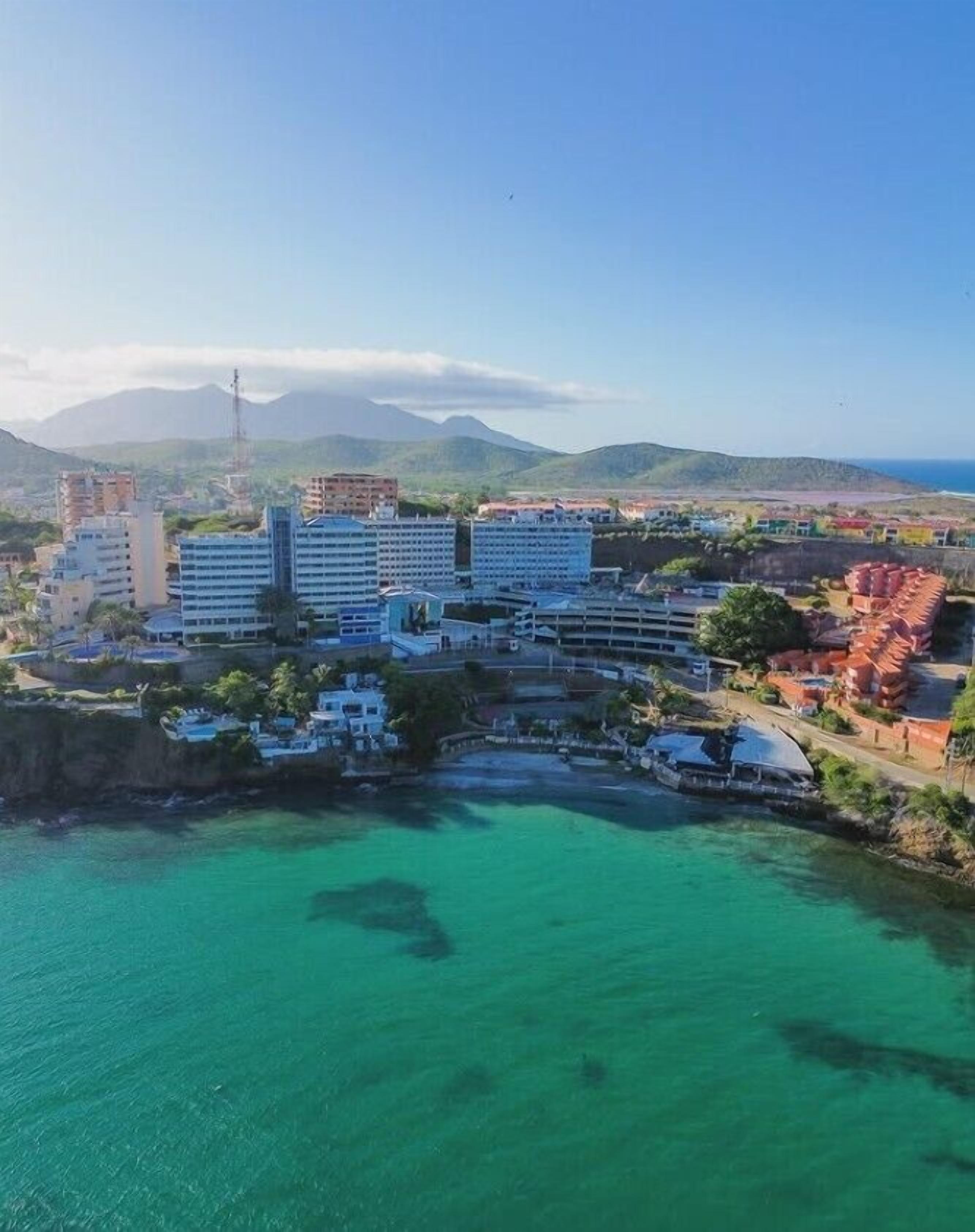 Vue aérienne d’une ville côtière, avec un imposant complexe hôtelier comprenant plusieurs bâtiments, un port de plaisance et des eaux turquoise d’une clarté remarquable.
