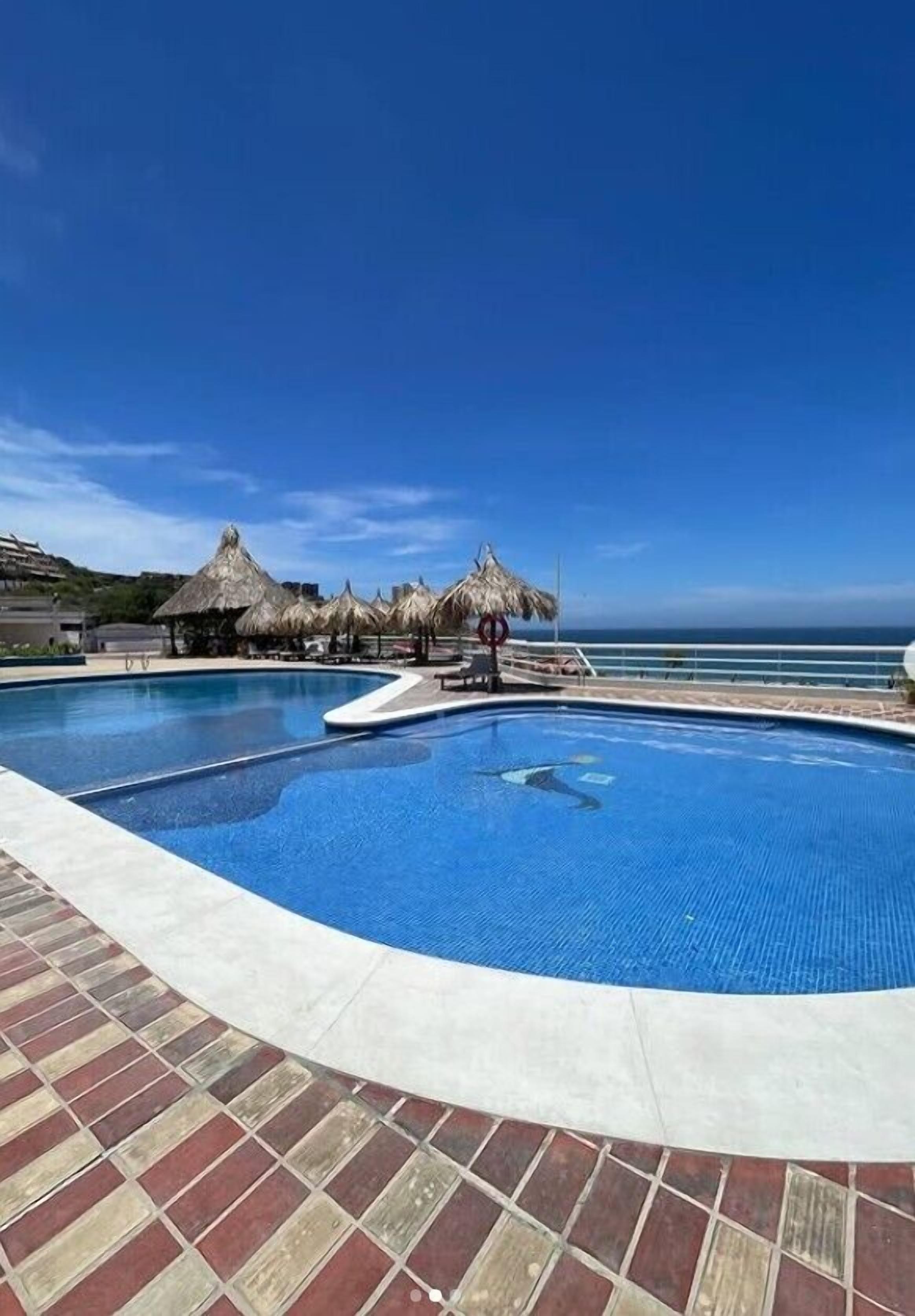 A swimming pool with a clear view of the ocean, surrounded by thatched umbrellas and a tiled deck.